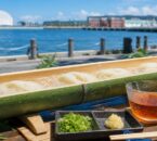港を背景に竹樋を流れる冷たいそうめんと、めんつゆ・薬味を並べた夏のイメージ