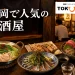 福岡の居酒屋で、もつ鍋、焼き鳥、刺身、ごまさば、日本酒が並ぶ夜の食卓を描いたアイキャッチ画像